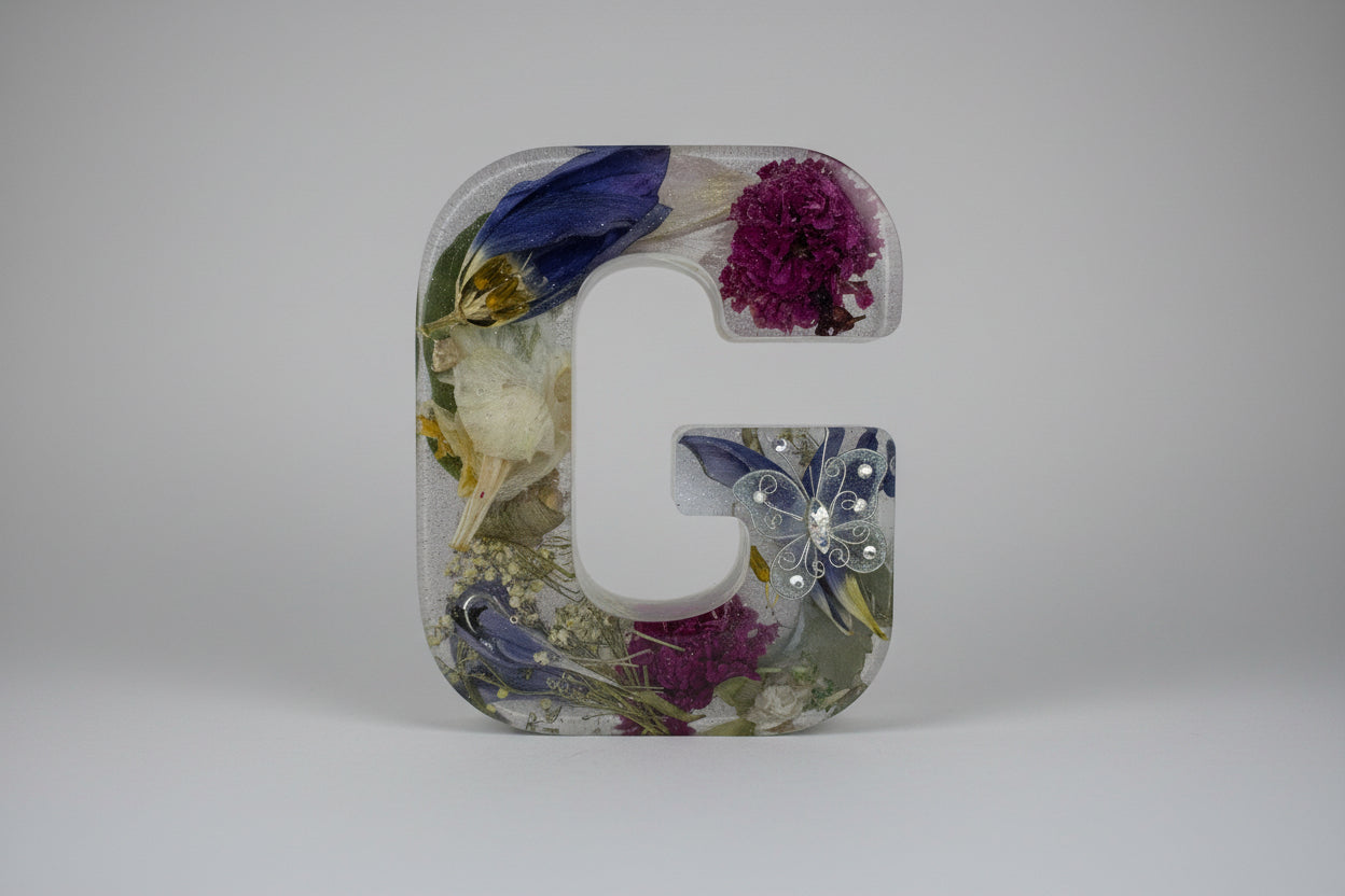 Hand holding a clear resin letter 'L' with embedded flowers and decorative elements on a white background.