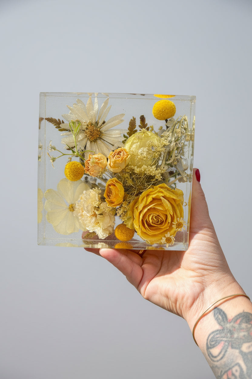 Hand holding a square transparent resin block with preserved flowers on a grey surface