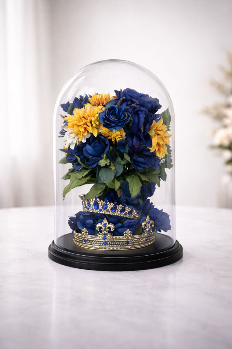 Decorative display with blue and yellow flowers and a crown under a glass dome on a white surface.