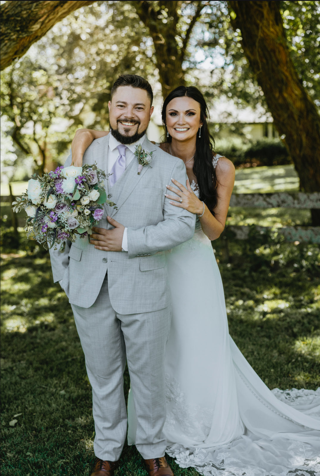 Couple in wedding attire standing under a tree with a bouquet of flowers.