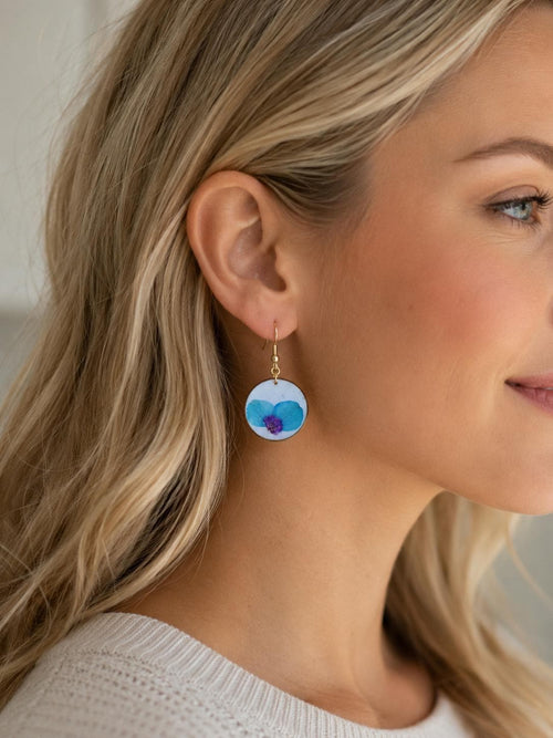 A close-up of a woman wearing a dangling round earring with a blue and purple watercolor-style design on a white background.