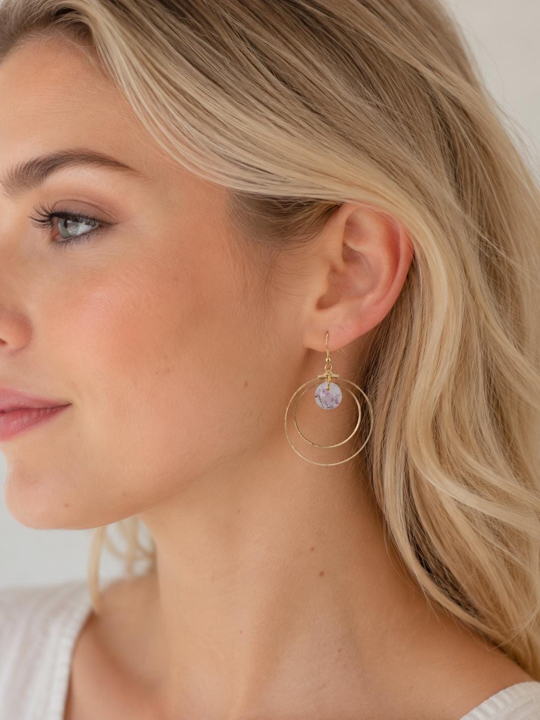 A close-up side profile of a blonde woman wearing a delicate gold hoop earring with a small round lavender-purple crystal drop.