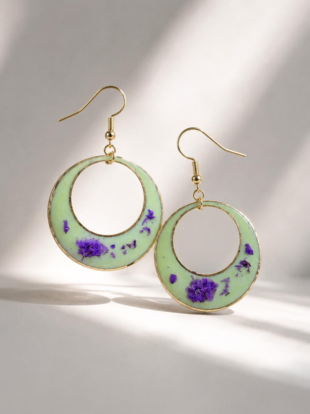Gold-tone circular drop earrings with a light green enamel-like center and embedded purple flower details, shown on a white background with soft diagonal shadows.