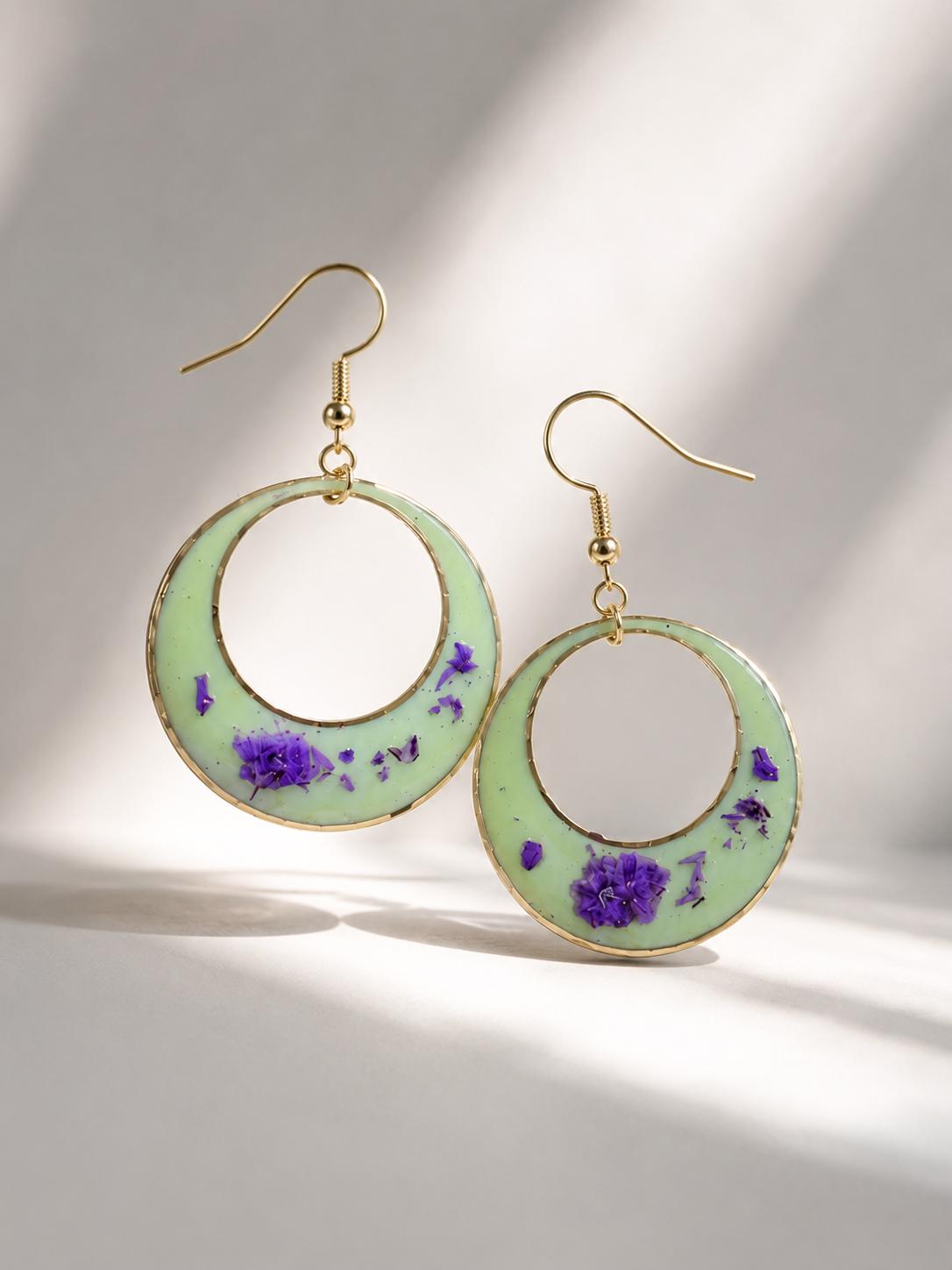 Gold-tone circular drop earrings with a light green enamel-like center and embedded purple flower details, shown on a white background with soft diagonal shadows.