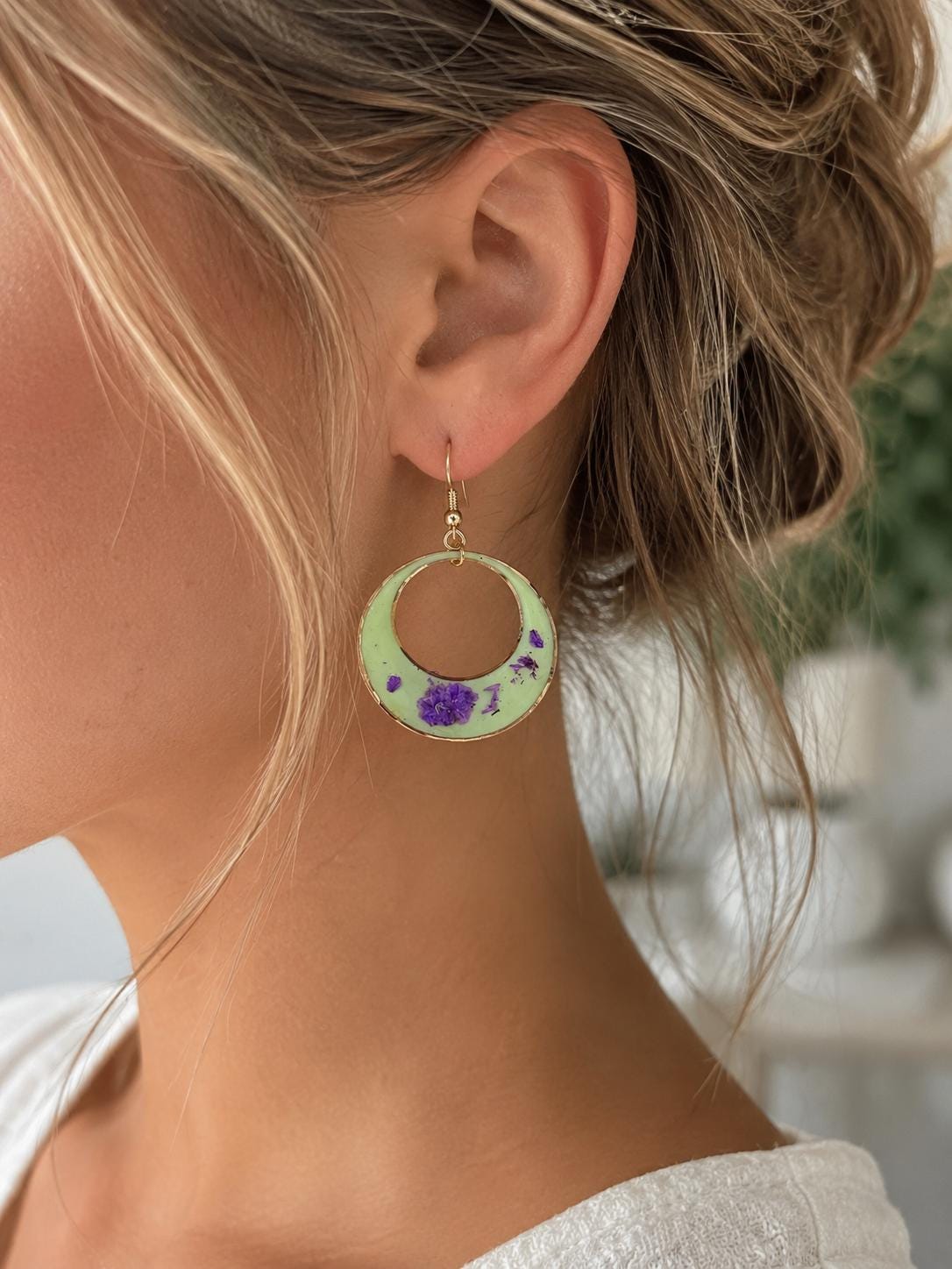 A close-up side profile of a woman wearing a dangling circular earring. The earring is light green with purple floral accents and a gold-tone hook.