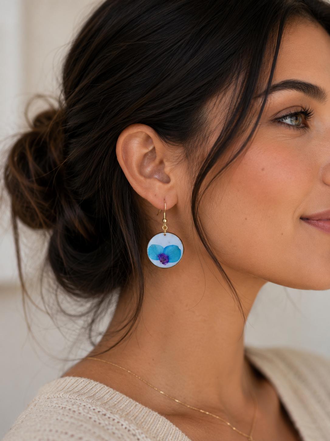 A close-up of a woman wearing a dangling earring with a round white top featuring a blue and purple flower design, suspended from a gold hook.