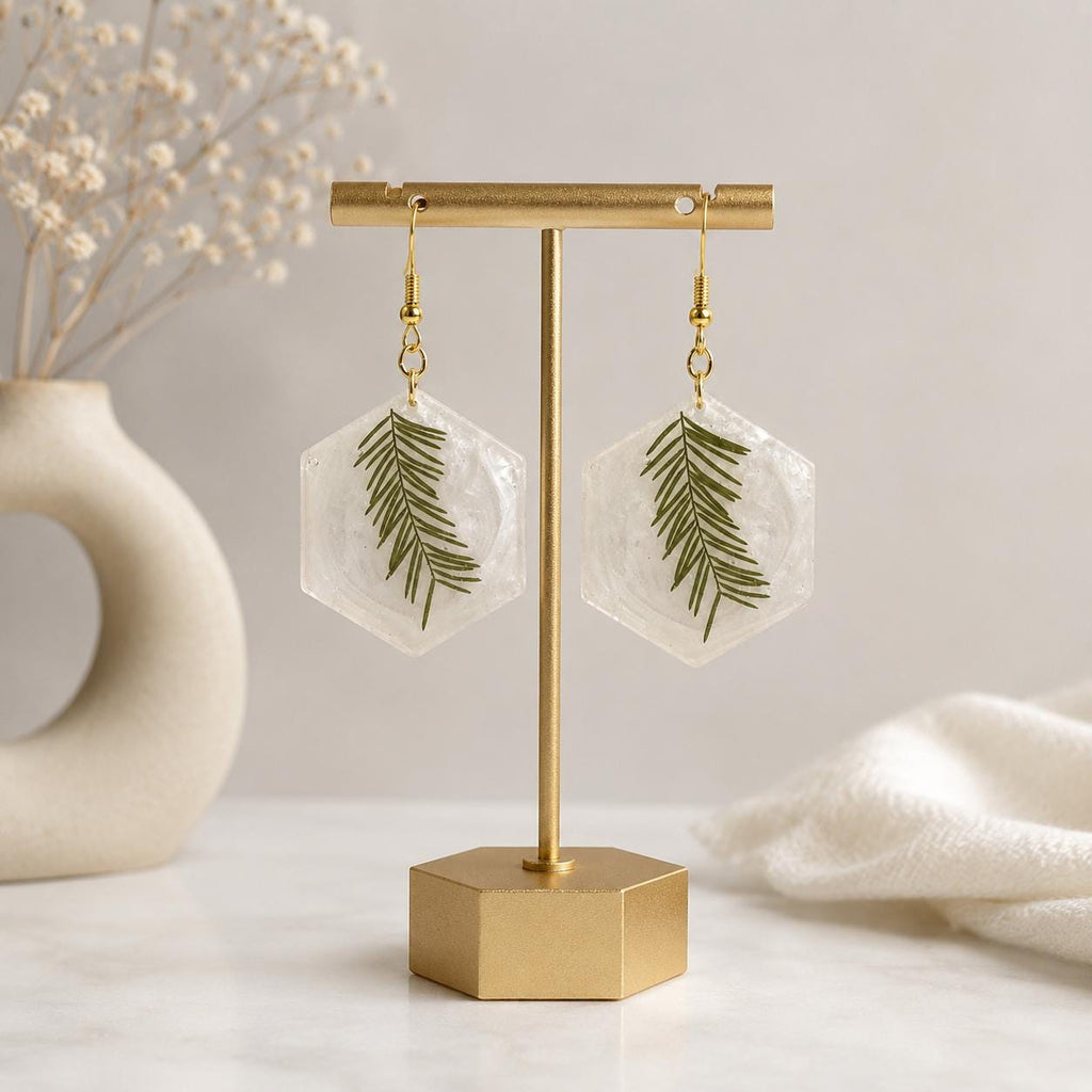 A pair of dangling earrings with pearlescent resin hexagonal pendants containing pressed green leaves, hanging from gold hooks on a gold T-bar jewelry display stand.