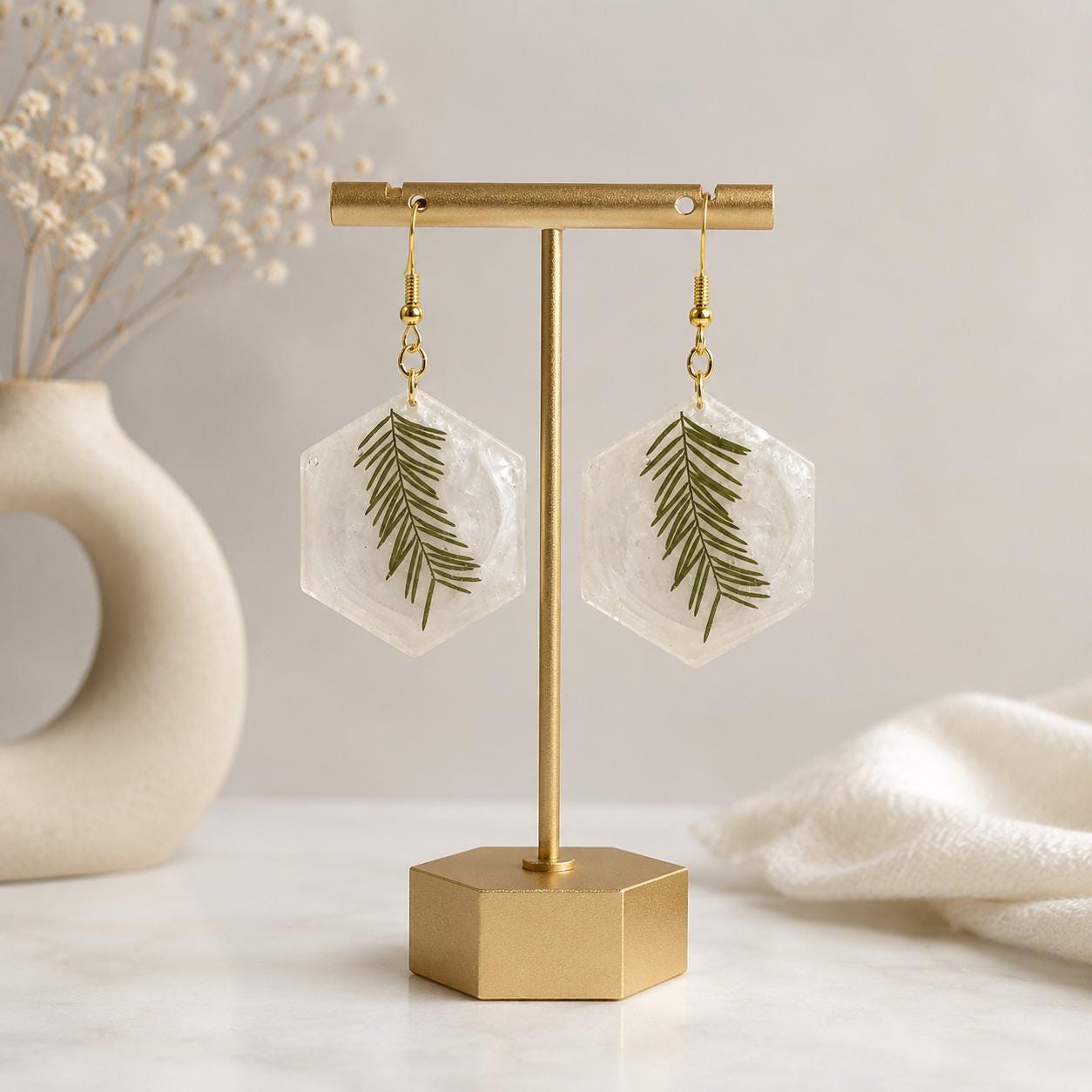 A pair of dangling earrings with pearlescent resin hexagonal pendants containing pressed green leaves, hanging from gold hooks on a gold T-bar jewelry display stand.