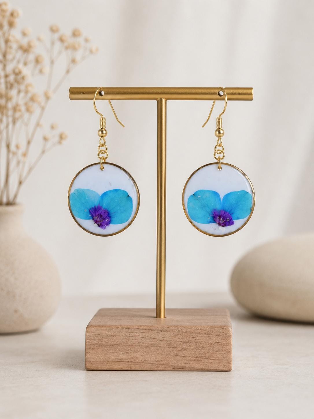 A pair of round gold-tone hoop earrings with embedded blue and purple flower petals, displayed on a gold T-stand with a wooden base against a soft beige background.