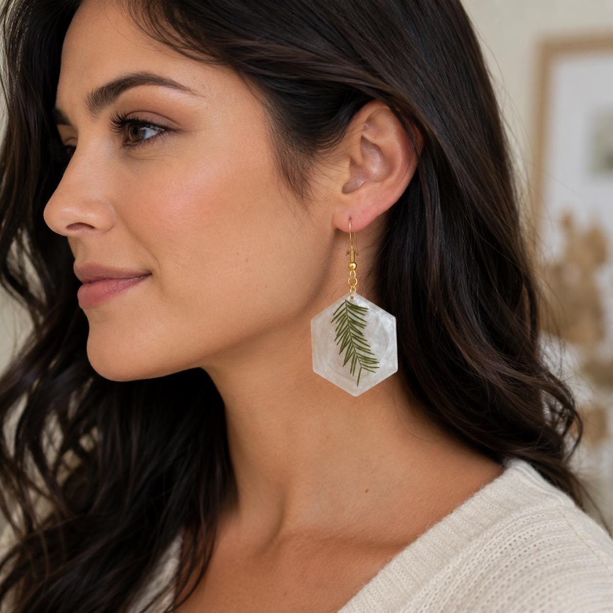 A woman in profile wearing a dangling hexagonal earring made of clear resin with a pressed green leaf and a small gold hook.