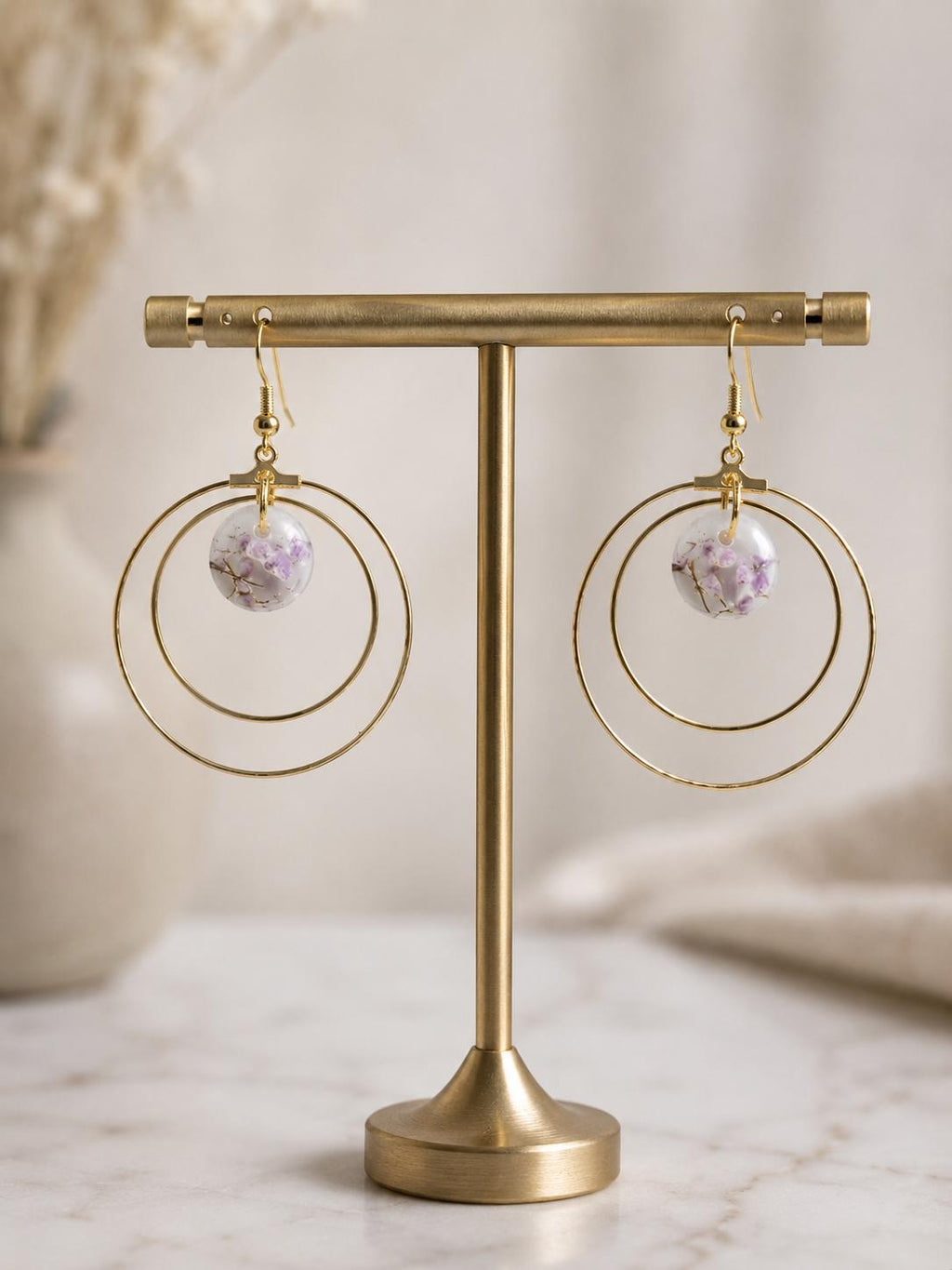 A pair of gold-tone hoop earrings with a small round lavender crystal pendant, displayed on a gold stand against a soft beige background.