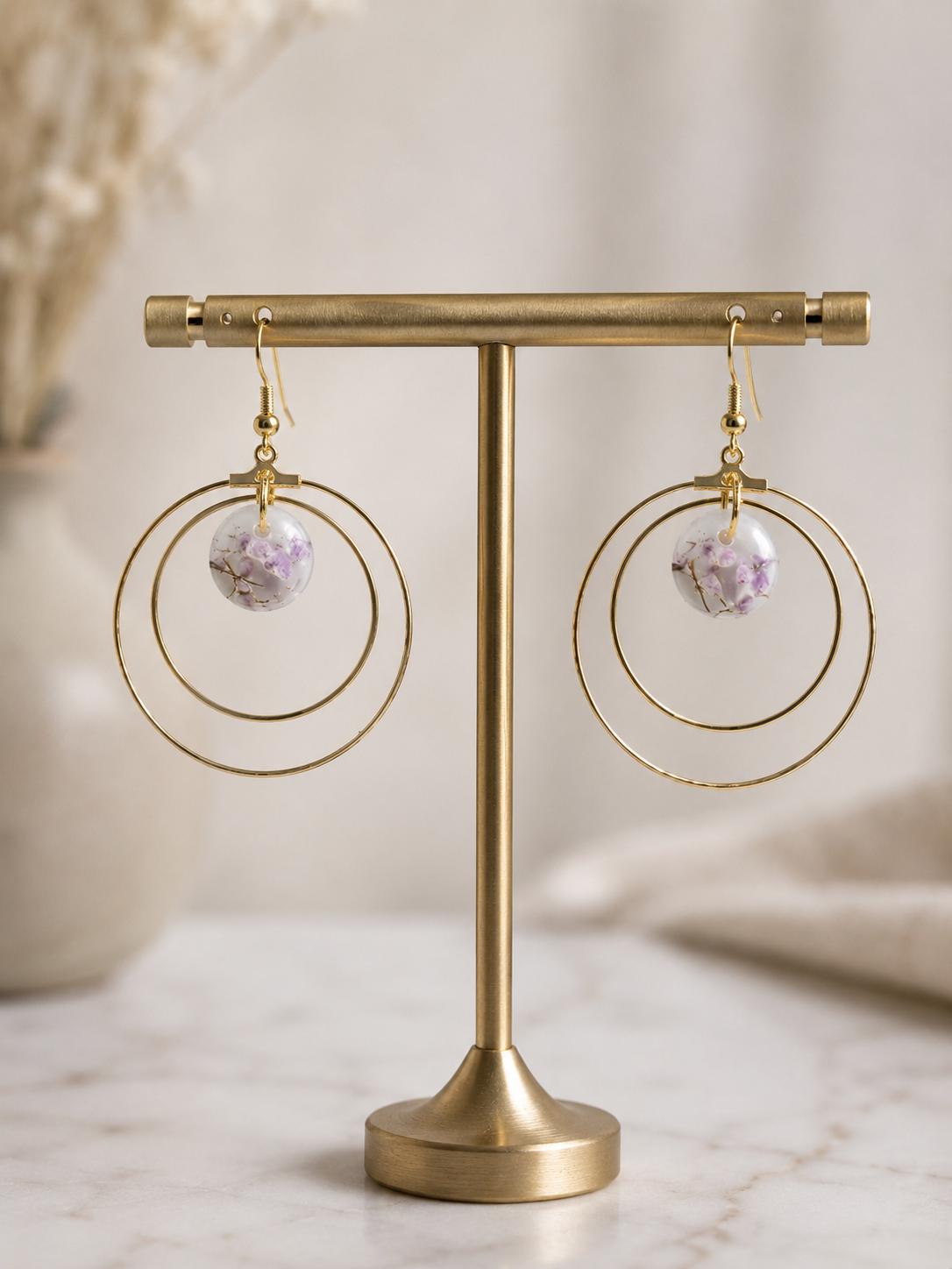 A pair of gold-tone hoop earrings with a small round lavender crystal pendant, displayed on a gold stand against a soft beige background.