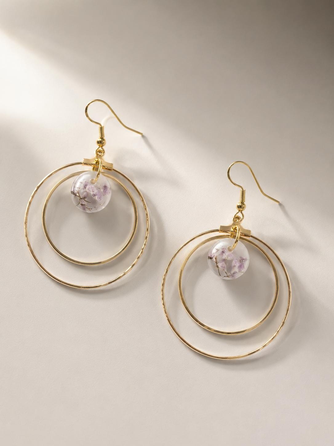 A pair of gold-toned double-ring hoop dangle earrings with a small faceted lavender crystal bead, displayed on a warm beige background with soft shadows.