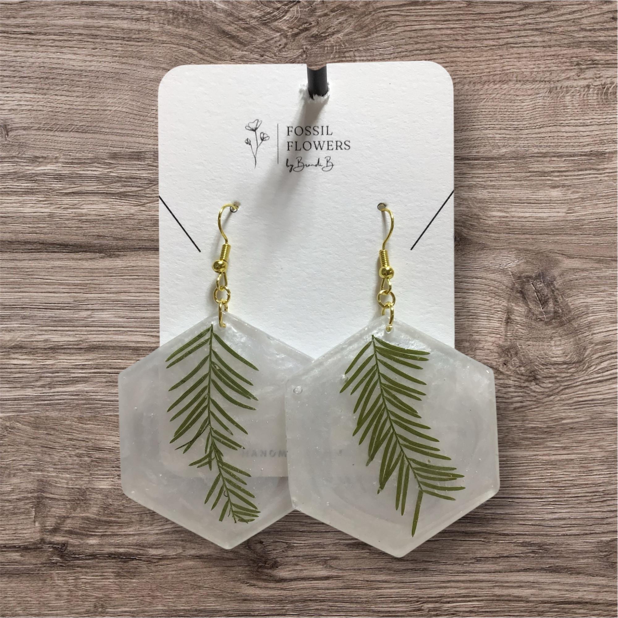 Hexagonal resin drop earrings with embedded green pine needles, hanging from gold-tone hooks on a white display card labeled Fossil Flowers, placed on a wood surface.