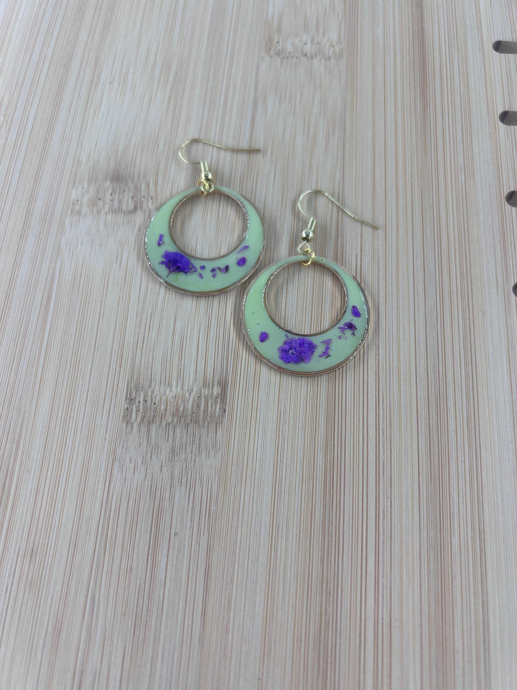 A pair of handmade crescent moon dangle earrings with a light green enamel finish, purple glitter accents, and gold-tone hooks, displayed on a light wood grain surface.