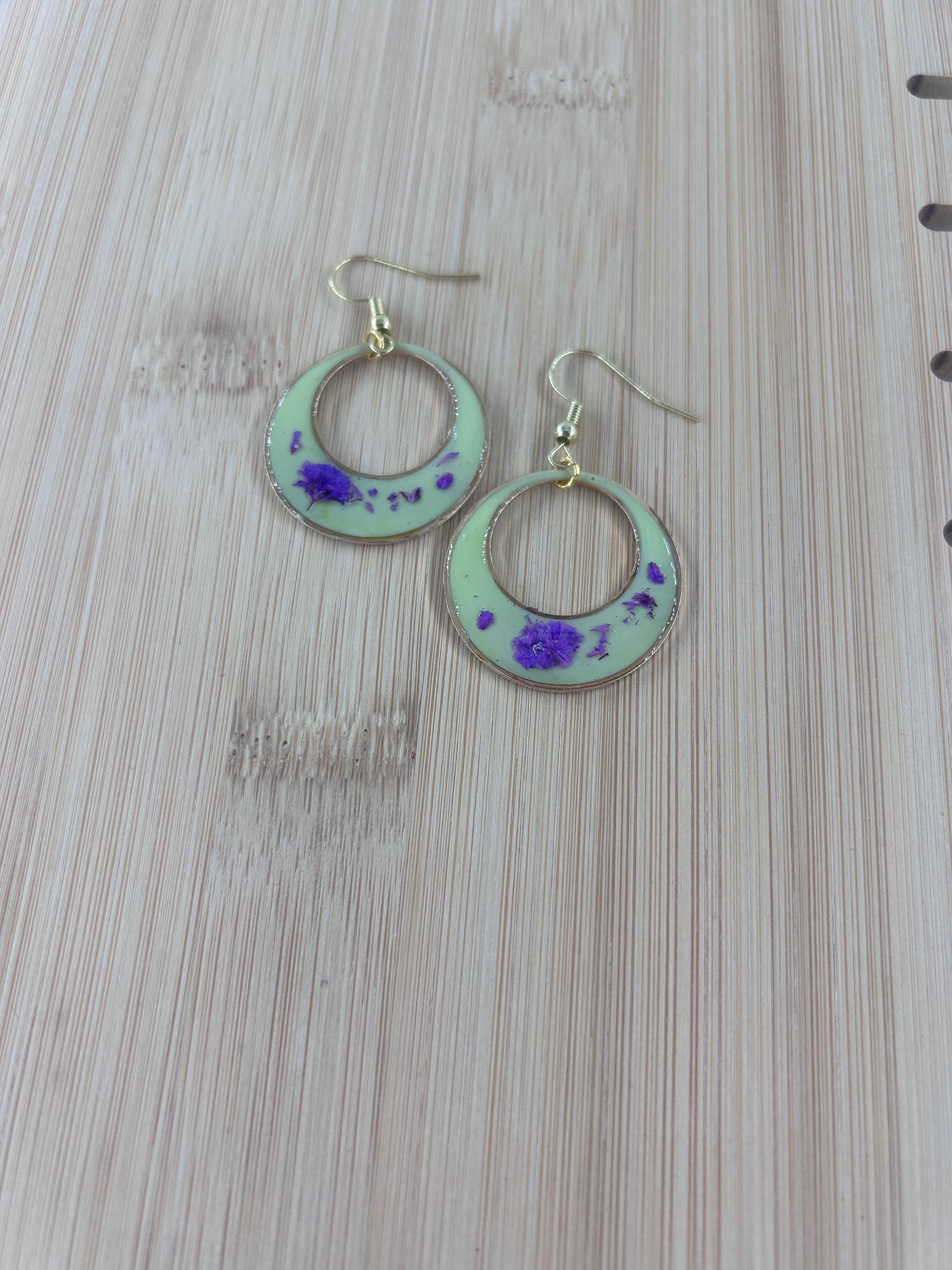 A pair of handmade crescent moon dangle earrings with a light green enamel finish, purple glitter accents, and gold-tone hooks, displayed on a light wood grain surface.