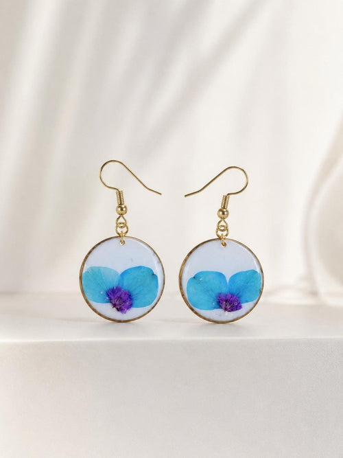 A pair of gold-tone dangle earrings with round white inlays featuring pressed blue flowers with purple centers, displayed on a clean white surface with soft draped fabric in the background.