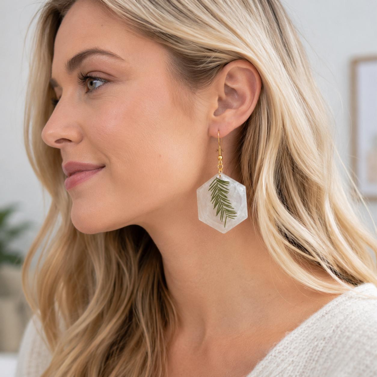 A close-up side profile of a woman with blonde hair wearing a dangling hexagonal earring made of clear resin with an embedded green leaf and a gold hook.