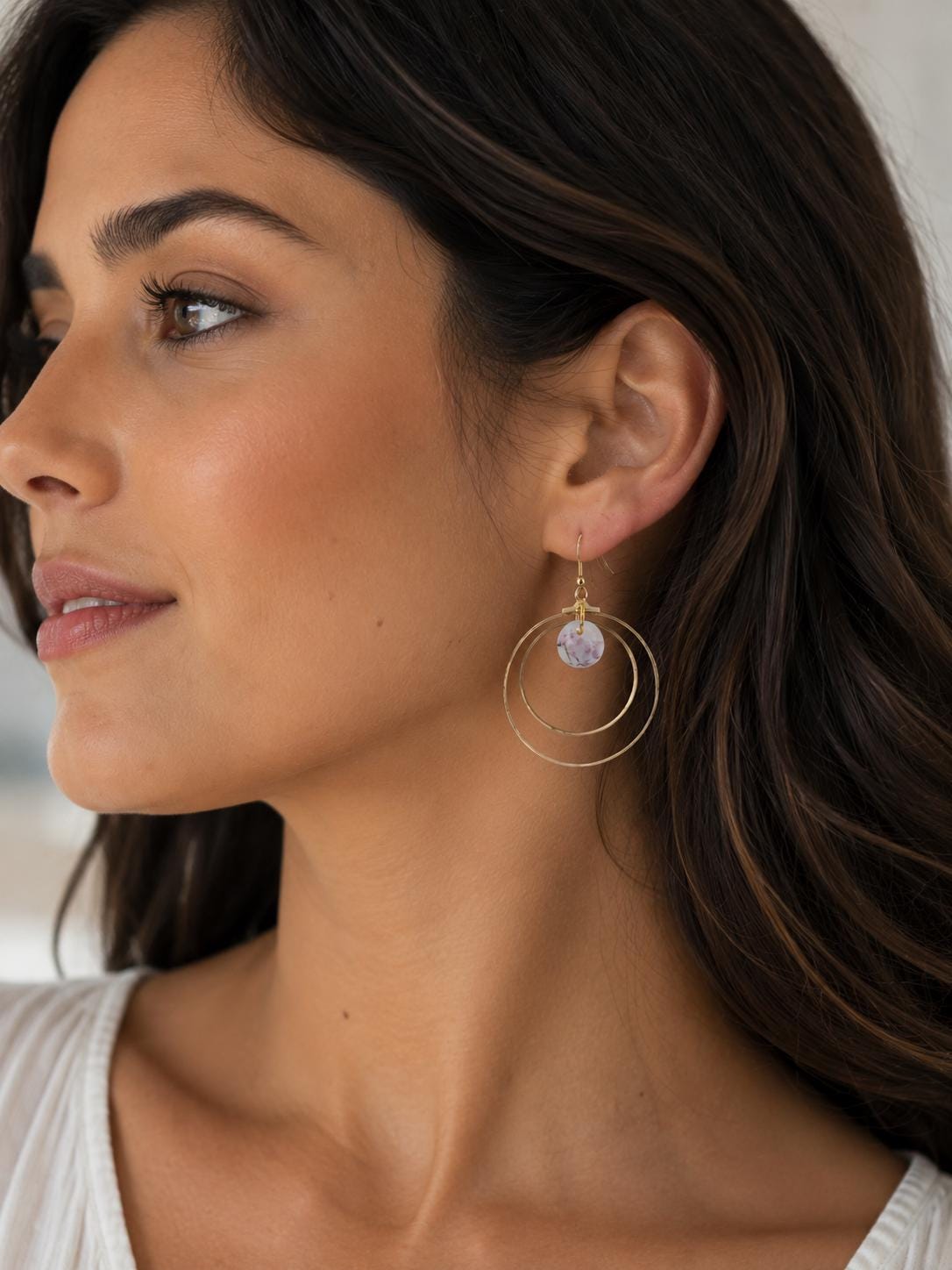A close-up side profile of a woman wearing a pair of gold-toned double-ring drop earrings with a small round lavender crystal charm.