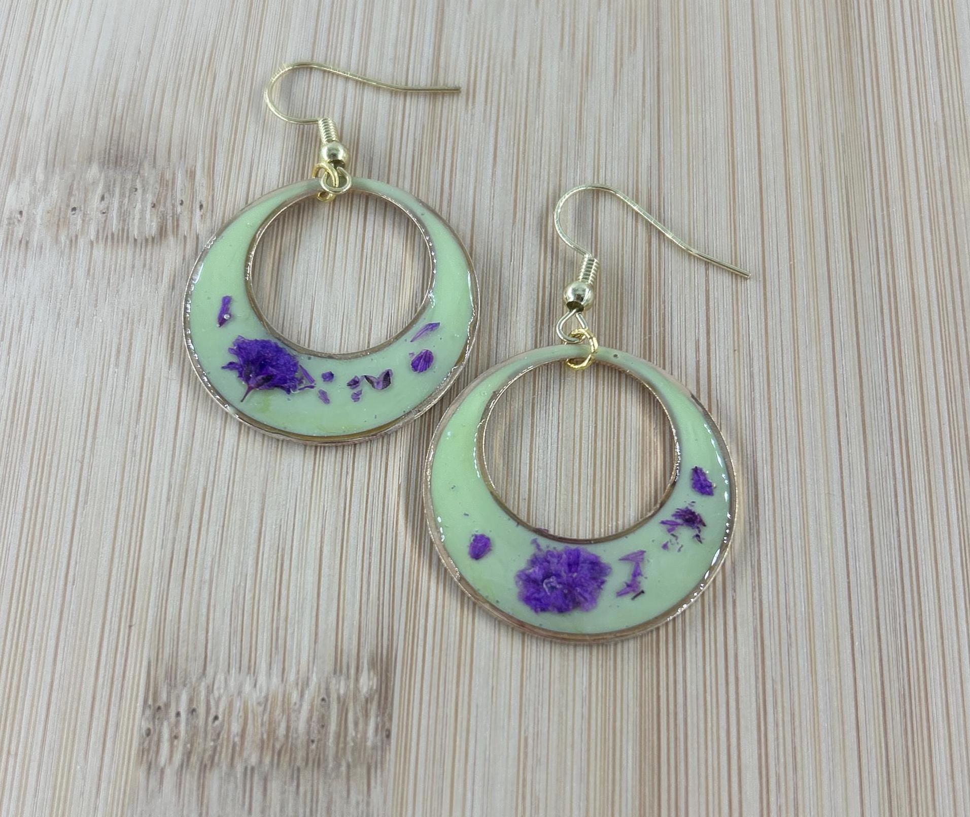 A pair of crescent-shaped hoop earrings with a pale mint green background, embedded with small purple dried flowers, and finished with gold-tone wire hooks, displayed on a light wood grain surface.