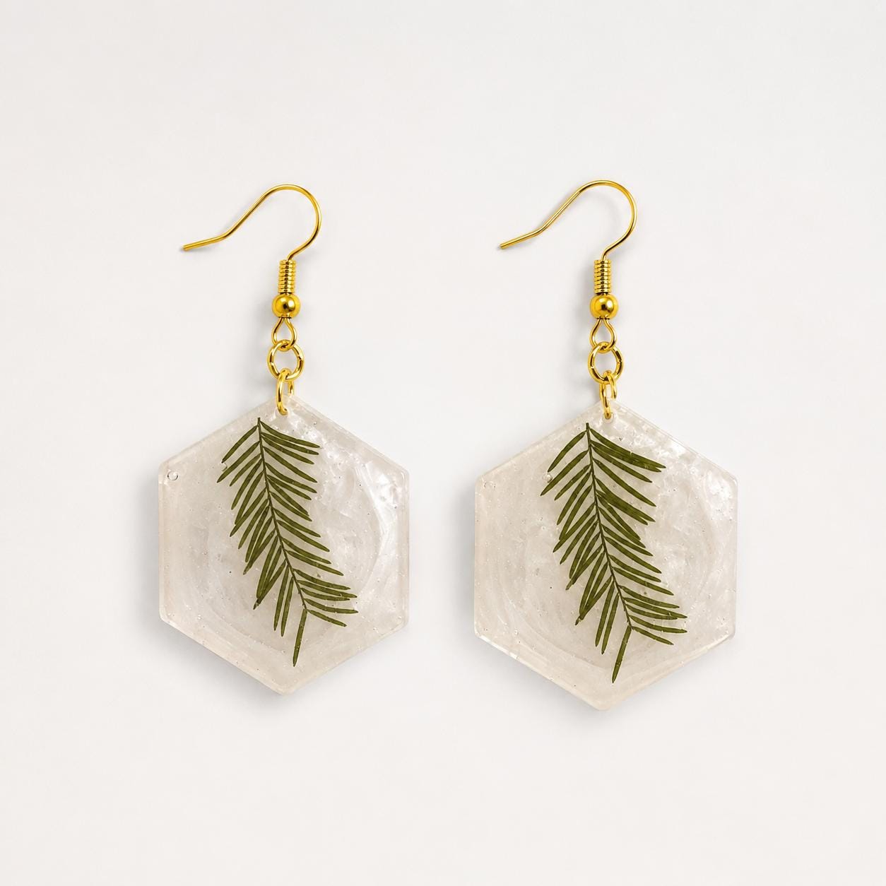 A pair of hexagonal dangle earrings with clear resin pendants containing green pine needles, suspended from gold-tone fishhook wires on a plain white background.