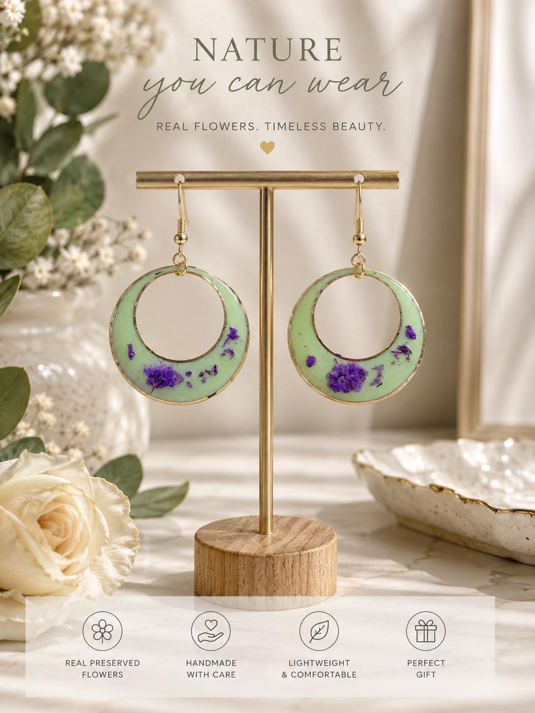 A pair of large circular hoop earrings displayed on a gold stand, featuring a pale mint green background with embedded purple flowers. The earrings are shown in a styled setting with flowers, a bowl, and the text 'NATURE you can wear' and 'REAL FLOWERS, TIMELESS BEAUTY.